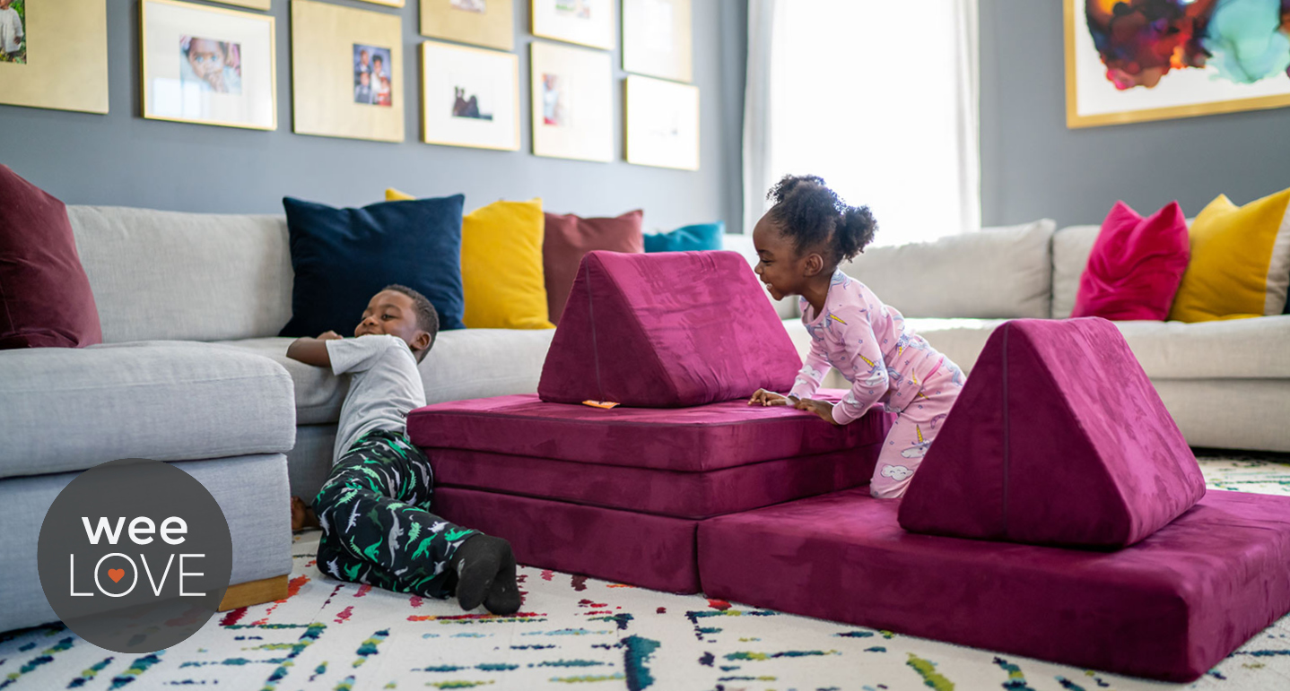 Two toddlers play on a magenta nugget couch that they have arranged on the floor of their living room like a pirate ship.