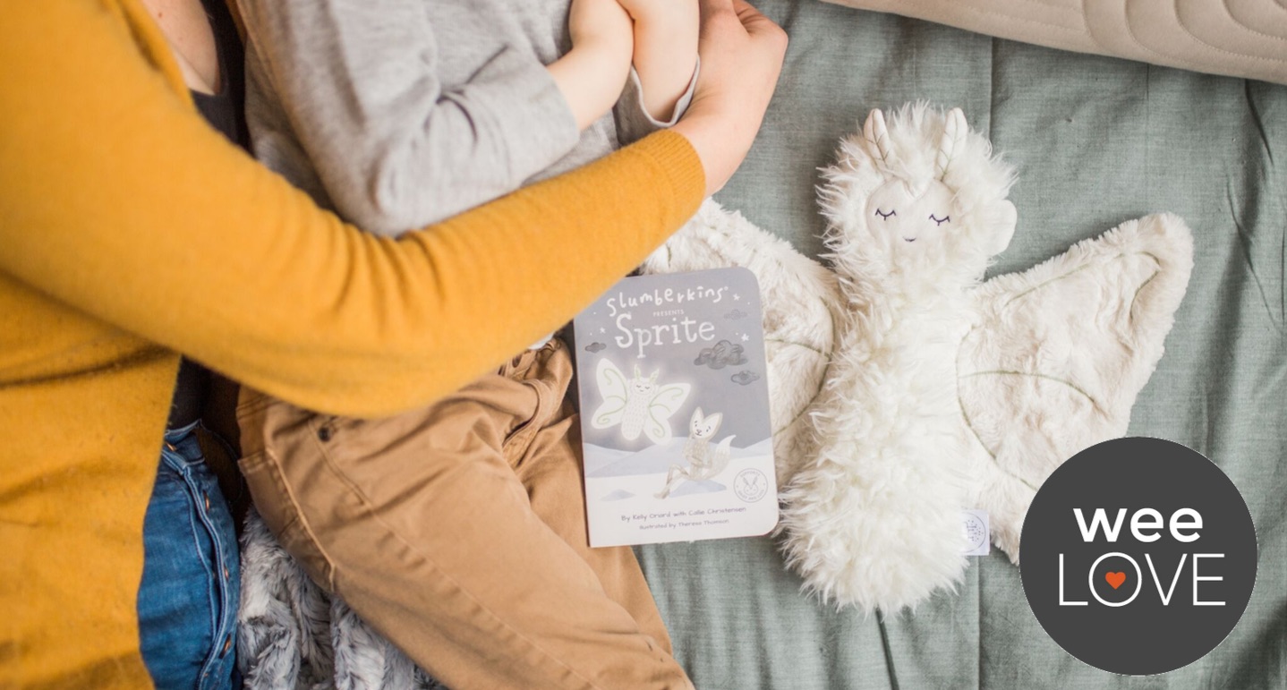 a parent hugs their child on a bed next to a slumberkins book and cuddle buddy, a moth named Sprite