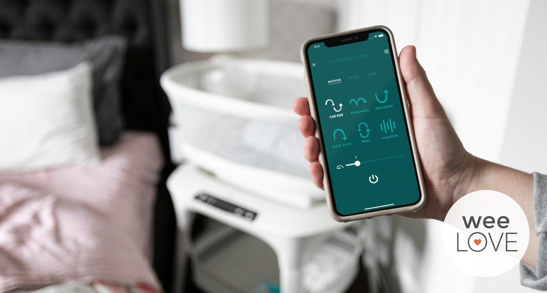 a hand holds a smart phone with the 4moms sleep bassinet app displayed on the screen, with the smart sleep bassinet in the background