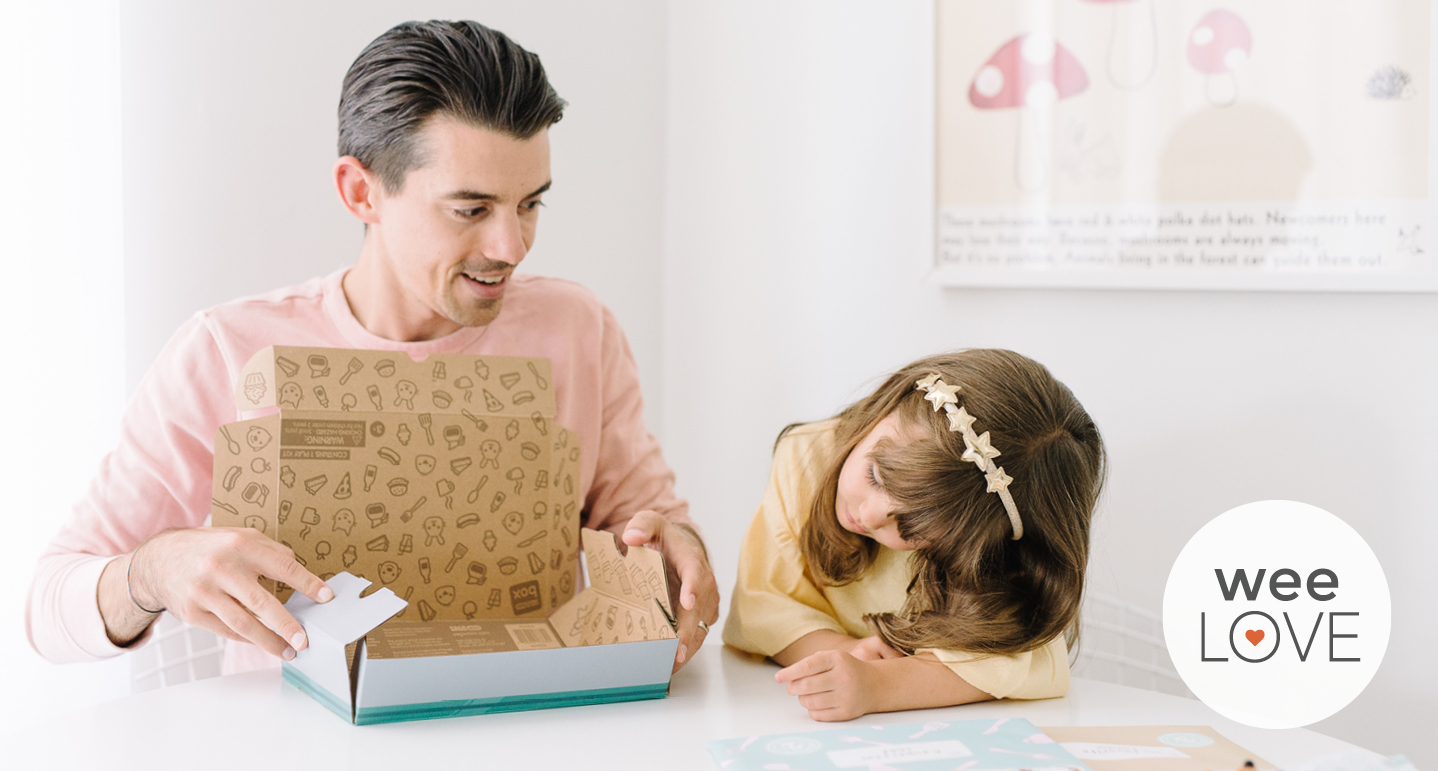 a father opens a Sago Mini subscription box on a kitchen counter while his preschool-aged daughter watches happily next to him