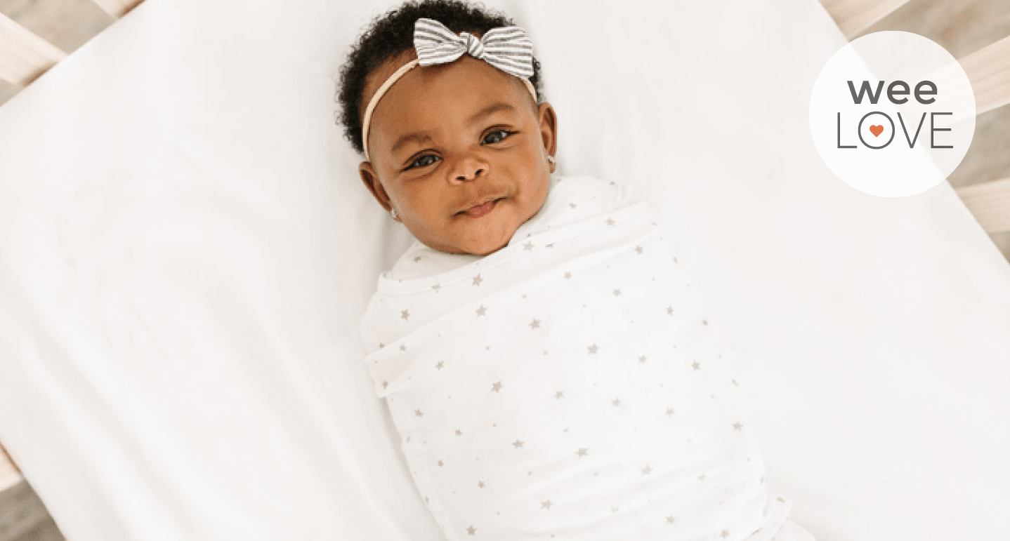 an aerial photo of a Black baby girl smiling up at the camera from her crib, wearing a dreamland baby swaddle.