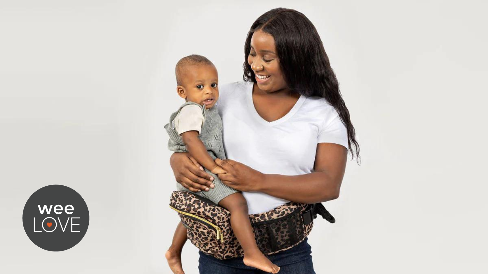 A Black mother in a white tshirt carries her Black, toddler-aged son a camo-printed Tushbaby, a hip carrier. She is smiling at her son, he is looking at the camera.
